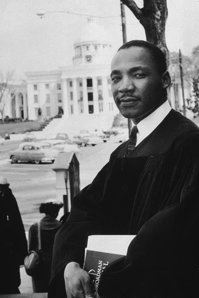 martin luther king jr looks at the camera while standing outside in a pastor robe over a collared shirt and tie, he holds papers in both hands in front of him, behind him is a street scene and a large white building