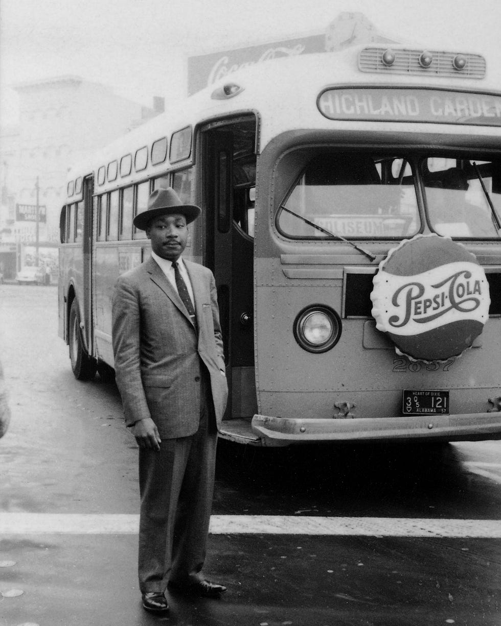 martin luther king jr stands outside in a suit and hat, behind him is a city bus with a pepsi cola ad on the front