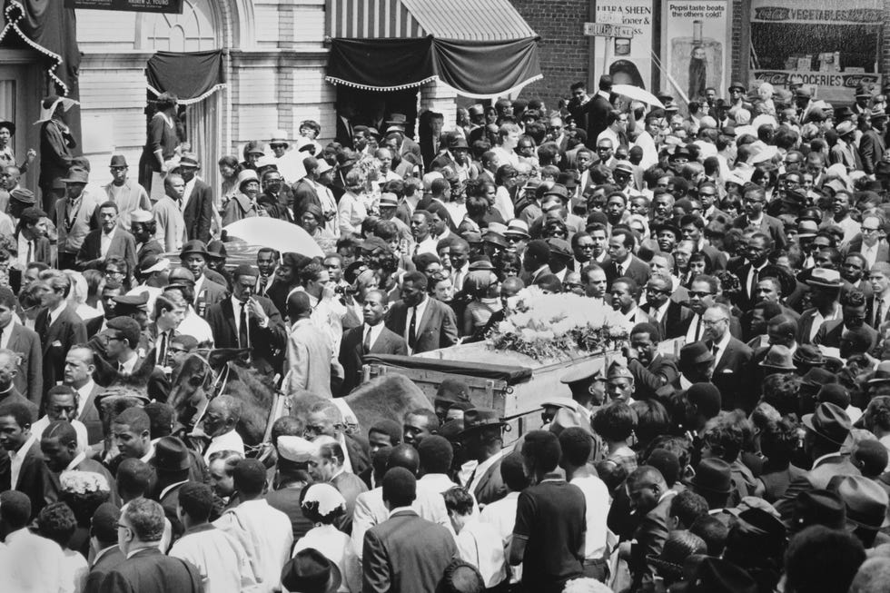 a crowd of people surround a horse drawn cart pulling a casket topped with flowers