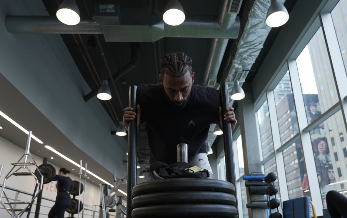 A man in a black t-shirt pushes a rack of weights in front of him in a gym setting. 